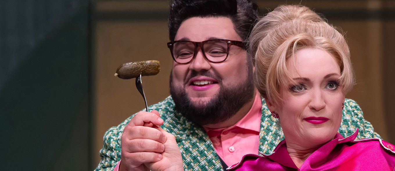 Szenenfoto aus der Operette "Messeschlager Gisela": Eine Frau hält einem Mann eine auf eine Gabel aufgespießte, saure Gurke unter die Nase.